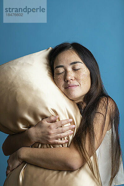 Woman with eyes closed hugging golden pillow against blue background