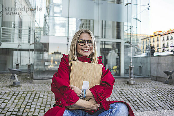 Glückliche reife Frau umarmt Laptop und sitzt auf Fußweg