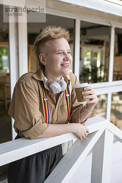 Lächelnde nicht-binäre Person mit Einwegkaffeetasse am Geländer