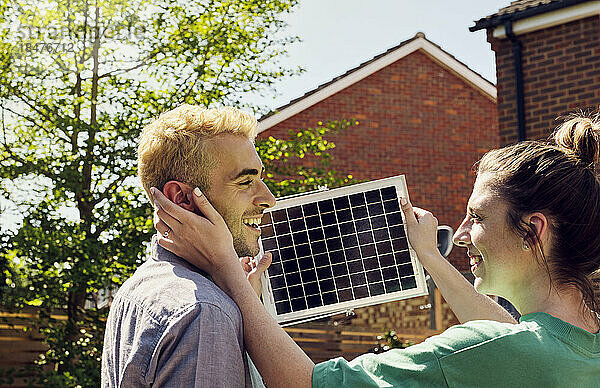 Glückliche junge Frau berührt Gesicht ihres Freundes  der Solarpanel hält
