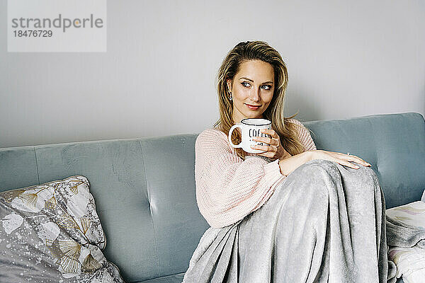 Nachdenklich lächelnde Frau sitzt mit Kaffeetasse auf dem Sofa zu Hause