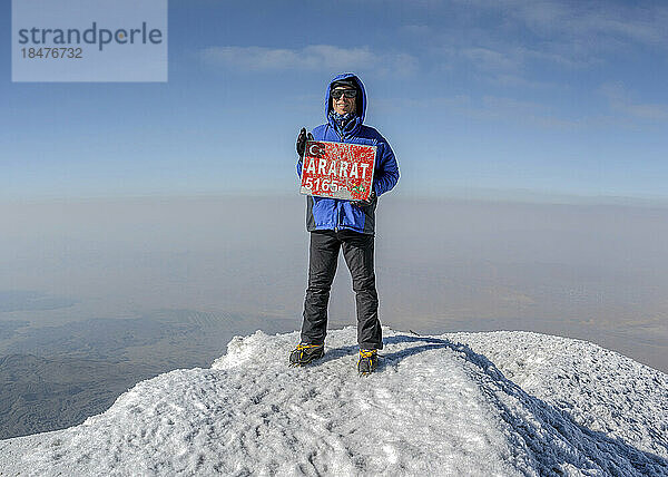 Lächelnder Wanderer mit Schild auf dem Gipfel des Berges Ararat an einem sonnigen Tag