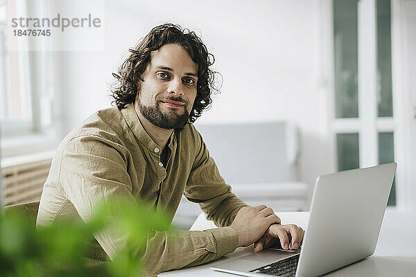 Lächelnder junger Geschäftsmann sitzt mit Laptop am Schreibtisch im Büro