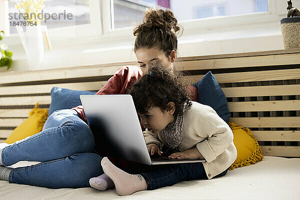 Kleines Mädchen sitzt mit Laptop auf dem Schoß der Mutter auf der Couch zu Hause