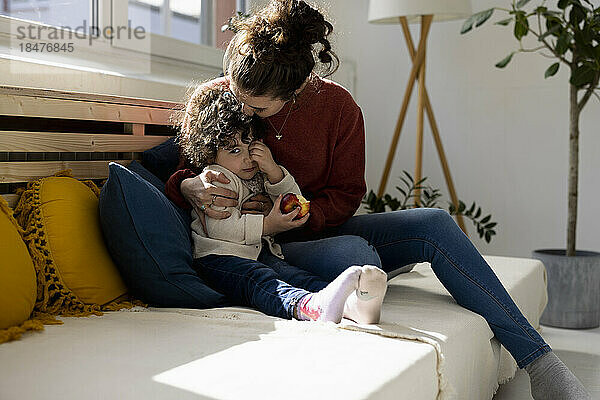 Mutter umarmt Tochter  die auf dem Sofa Apfel isst