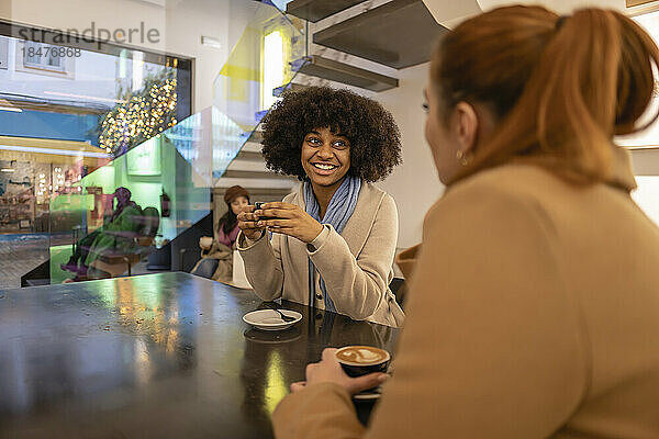 Glückliche Frau hält Kaffeetasse in der Hand und spricht mit einer Freundin im Café