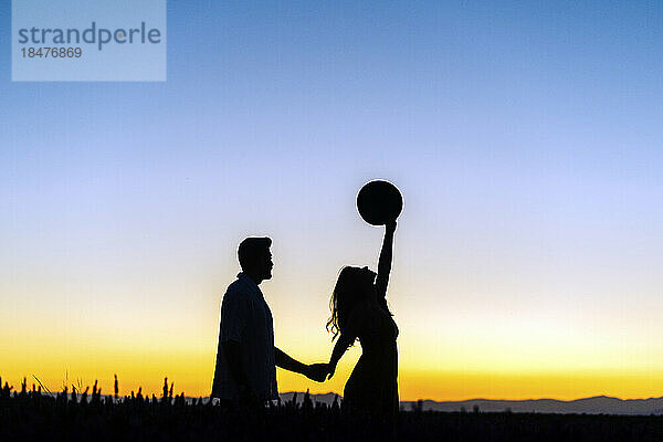 Silhouette eines Mannes  der seine Freundin an der Hand hält und bei Sonnenuntergang den Hut im Feld hebt