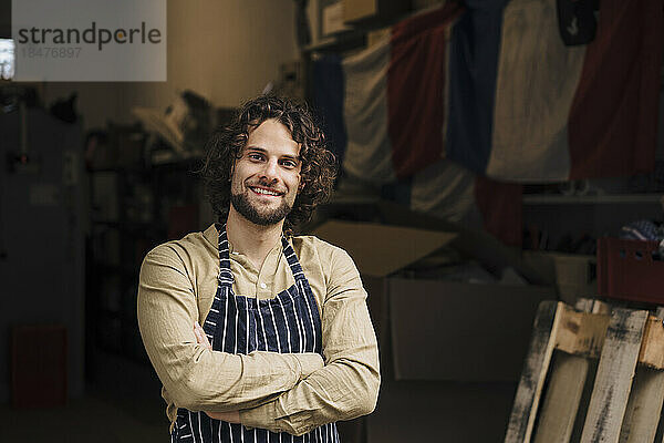 Happy young businessman wearing apron standing with arms crossed at storage room