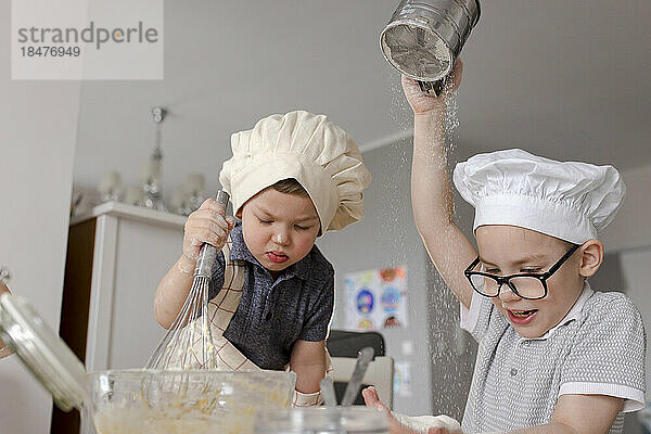 Boy sieving flour by brother mixing batter with whisk in kitchen