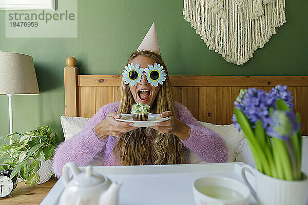 Fröhliche Frau mit Sonnenblumen-Sonnenbrille hält zu Hause im Bett einen Teller mit Cupcake