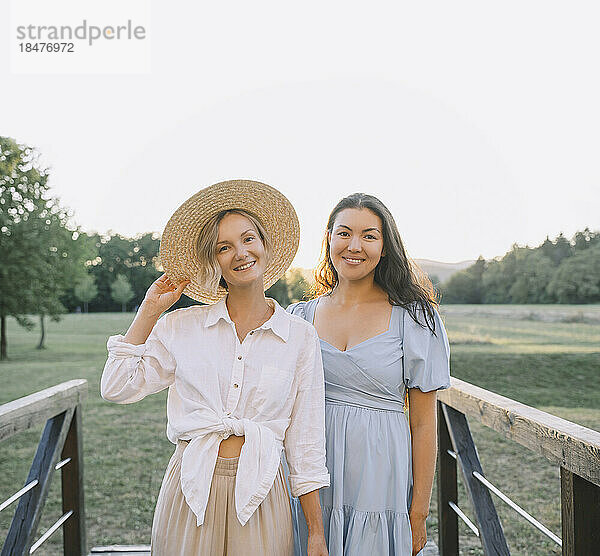 Lächelnde Frau mit Hut steht mit Freundin am Geländer