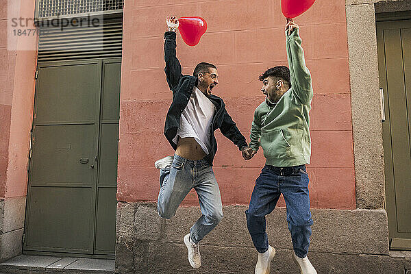 Glückliches schwules Paar  das mit roten herzförmigen Luftballons springt