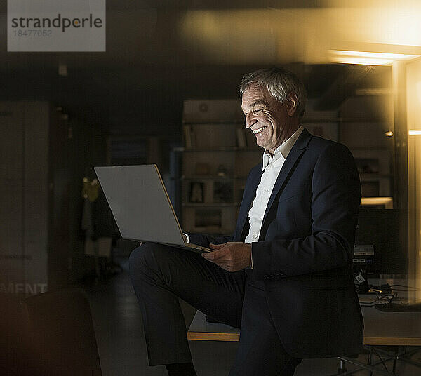Glücklicher Geschäftsmann mit Laptop  der im Büro sitzt