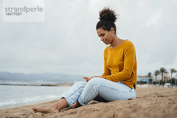 Junge Frau benutzt Mobiltelefon und sitzt auf Sand