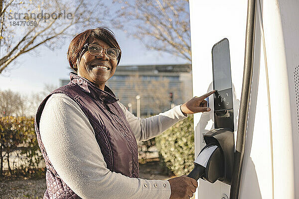 Lächelnde  nachdenkliche ältere Frau  die an der Elektroladestation steht