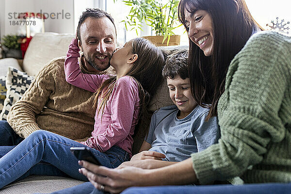 Glückliche Mutter benutzt Smartphone  während Sohn und Mädchen zu Hause den Vater küssen