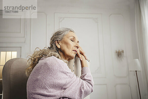 Ältere Frau mit geschlossenen Augen sitzt zu Hause auf einem Stuhl