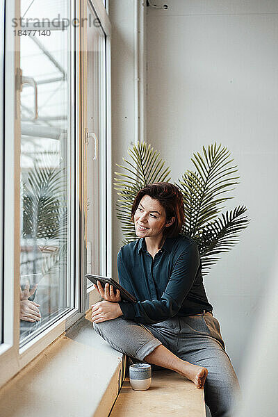 Lächelnde junge Geschäftsfrau mit Tablet-PC  die durch das Fenster im Büro schaut
