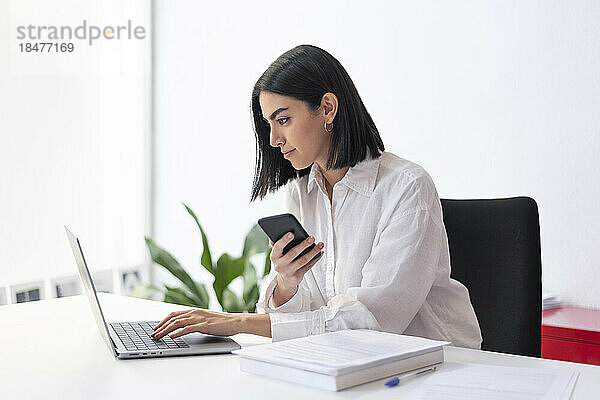 Junge Geschäftsfrau mit Smartphone und Laptop am Schreibtisch