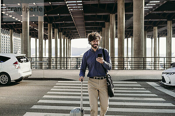 Glücklicher Geschäftsmann mit Gepäck und Smartphone auf der Straße