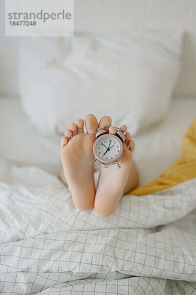 Feet of girl with alarm clock on bed at home