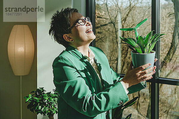 Fröhliche reife Geschäftsfrau mit Windturbinenmodell in Topfpflanze am Fenster im Büro