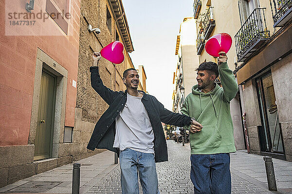 Fröhliches schwules Paar  das sich mit roten herzförmigen Luftballons inmitten von Gebäuden auf der Straße amüsiert