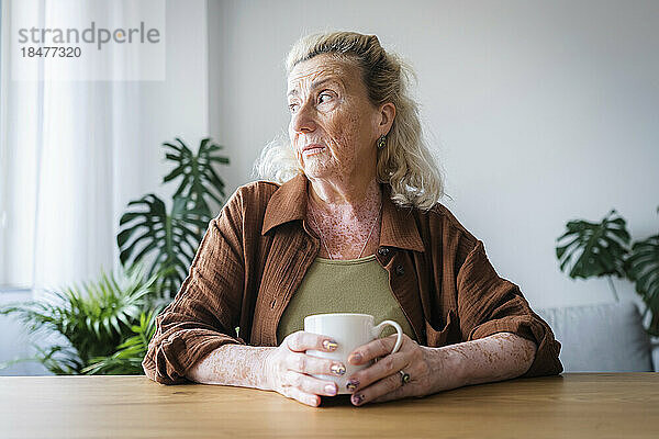 Besorgte ältere Frau hält einen Becher in der Hand und sitzt zu Hause