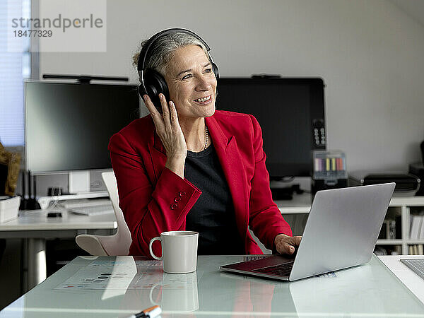 Glückliche ältere Geschäftsfrau mit kabellosen Kopfhörern sitzt mit Laptop am Schreibtisch
