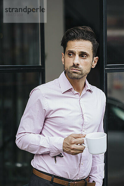 Nachdenklicher Geschäftsmann steht mit Kaffeetasse vor der Tür