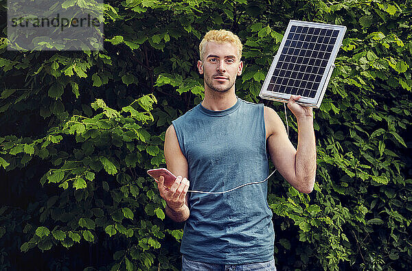 Junger Mann lädt Smartphone über Solarpanel vor Pflanzen auf