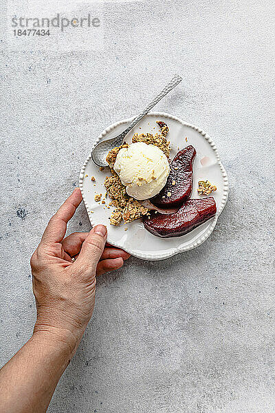 Woman's hand holding plate of pears in red wine with ice cream and granola