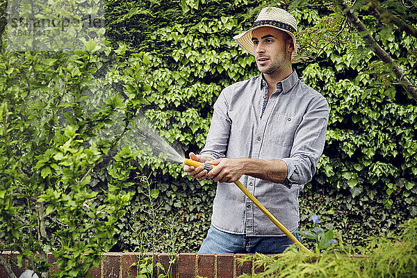 Junger Mann mit Hut gießt Pflanzen mit Schlauch