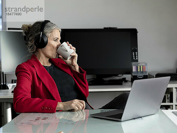 Ältere Geschäftsfrau trägt kabellose Kopfhörer und trinkt Kaffee im Büro