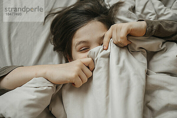 Verspielter Junge bedeckt sein Gesicht mit einer Decke auf dem Bett zu Hause