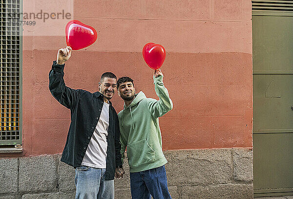 Glückliches junges schwules Paar hält roten Ballon vor der Wand