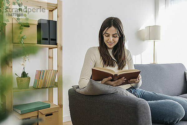 Lächelnde Frau  die auf dem Sofa im Wohnzimmer ein Buch liest