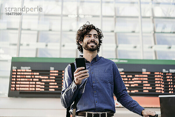 Glücklicher Geschäftsmann mit Smartphone  der vor der Ankunfts- und Abflugtafel steht