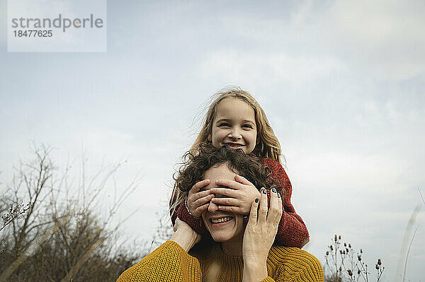 Lächelndes Mädchen  das die Augen seiner Mutter vor dem Himmel bedeckt