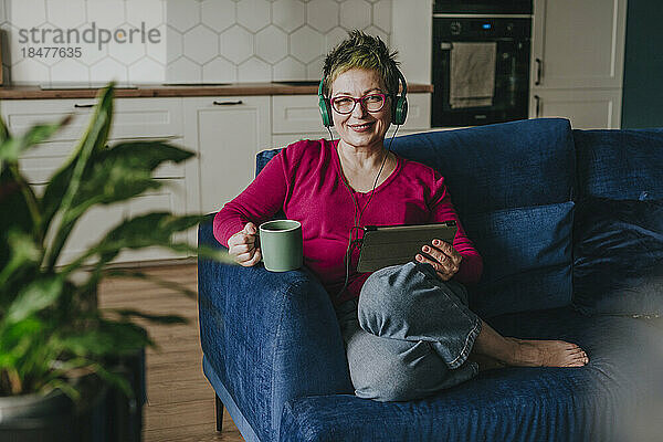 Smiling woman wearing headphones sitting with tablet PC on sofa at home