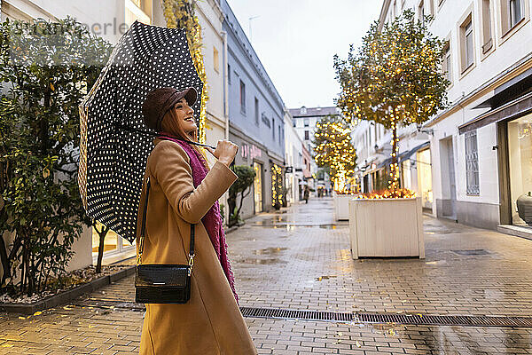 Lächelnde junge Frau mit Regenschirm steht am Fußweg