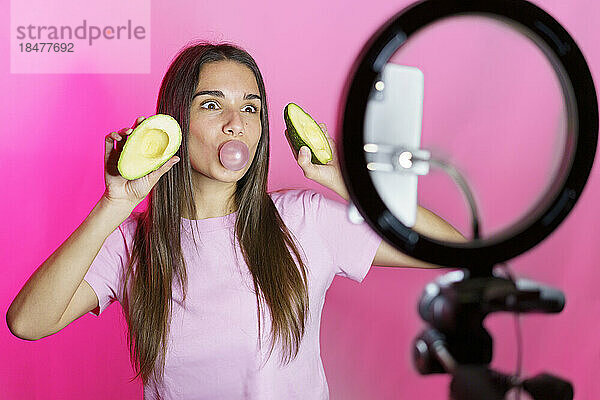 Frau mit Avocadohälften bläst Seifenblasen und vloggt vor rosa Hintergrund