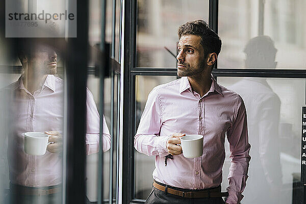 Nachdenklicher Geschäftsmann hält Kaffeetasse vor Glaswand