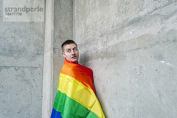 Nicht-binäre Person  eingehüllt in eine Regenbogenfahne  steht an der Wand