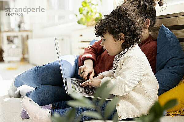 Kleines Mädchen sitzt mit Laptop auf dem Schoß der Mutter auf der Couch zu Hause