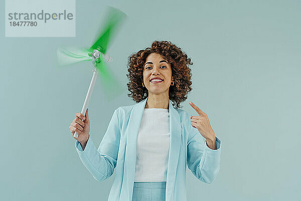 Happy woman gesturing at spinning wind turbine model against blue background