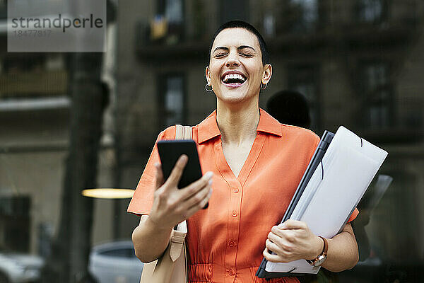 Geschäftsfrau mit Mobiltelefon und Akten lacht vor Glaswand