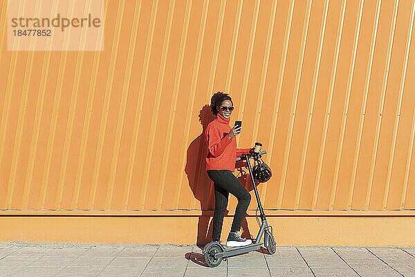 Frau benutzt Smartphone mit Elektroroller vor orangefarbener Wand