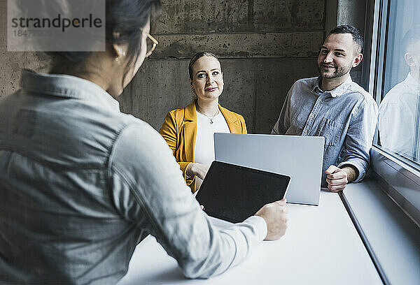 Lächelnde Kollegen schauen einer Geschäftsfrau zu  die bei einer Besprechung im Büro einen Tablet-PC benutzt
