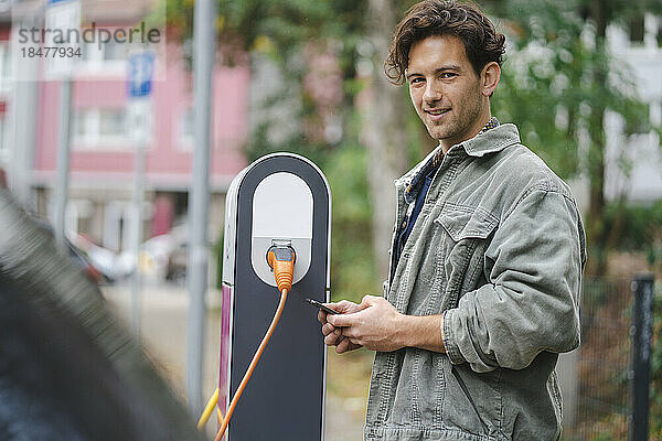 Lächelnder Mann steht mit Smartphone an der Ladestation für Elektrofahrzeuge